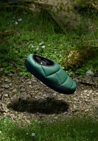 Green quilted slipper with black sole and interior, featuring a pull loop. Surrounded by grass and small flowers on a rocky surface.