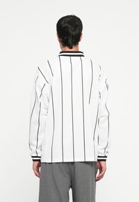 Chemise blanche à manches longues avec des rayures verticales noires, dotée d'un col noir et de panneaux latéraux texturés, portée avec un pantalon gris.