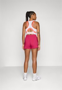 Top desportivo branco com detalhes em néon e costas abertas; calções rosa até acima do joelho; ténis brancos com solas cinzentas; superfície lisa.