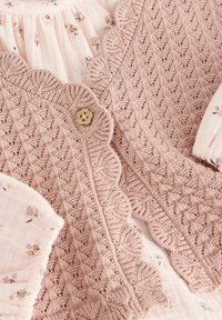 Lichtroze gebreide cardigan met geschulpte randen, voorzien van een bloemenknop en een ingewikkeld textuurpatroon; gecombineerd met een bloes met bloemen.