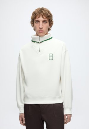 Junger Mann mit lockigem Haar, der einen weißen Pullover mit grün gestreiftem Reißverschlusskragen und kleinem Patch-Logo auf der Brust trägt.
