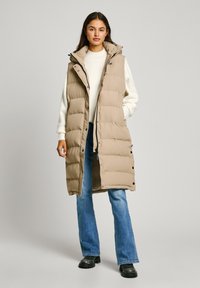 Beige pufferväst med hög krage, tryckknappar och kviltad design. Bärs över en krämfärgad tröja, i kombination med utsvängda jeans och svarta stövlar.