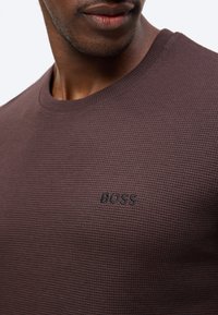 Κοντινό πλάνο του κάτω μέρους του προσώπου και του κορμού ενός άνδρα που φοράει καφέ υφασμάτινο πουλόβερ με κεντημένη τη λέξη "BOSS" στο στήθος.