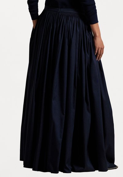 Sieviete ģērbusies garā, plūstošā tumši zilā svārkā ar krokām, kas sader ar tādu pašu tumši zilo garo piedurkņu blūzi, rokas atpūtina blakus.