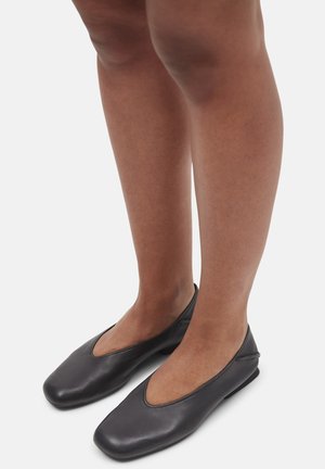 Jambes nues portant des ballerines noires en cuir à enfiler avec des orteils carrés arrondis, debout sur une surface blanche.
