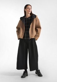 Bruine oversized jas met een zwarte kraag, gedragen over een zwart-wit geblokte trui en losse zwarte broeken. Zwarte sneakers.