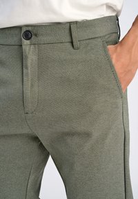 Olivgrüne Hose aus strukturiertem Stoff, mit taillierter Passform, Fronttaschen und Knopfverschluss. Vertikale Nähte sorgen für subtile Details.