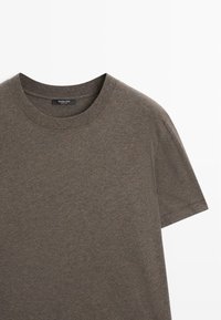 T-shirt in cotone marrone con scollo rotondo e maniche corte. Presenta una texture morbida e un design minimalista. Include un'etichetta sul collo.