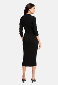 Robe noire en maille côtelée avec une silhouette ajustée, longueur midi, manches longues et col. Soulignée par une texture subtile.