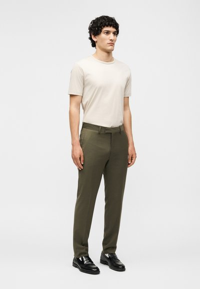 Νέος άνδρας που στέκεται με το πρόσωπο προς τα εμπρός, φοράει μπεζ κοντομάνικο πουκάμισο, χακί παντελόνια και μαύρα δερμάτινα παπούτσια σε λευκό φόντο.