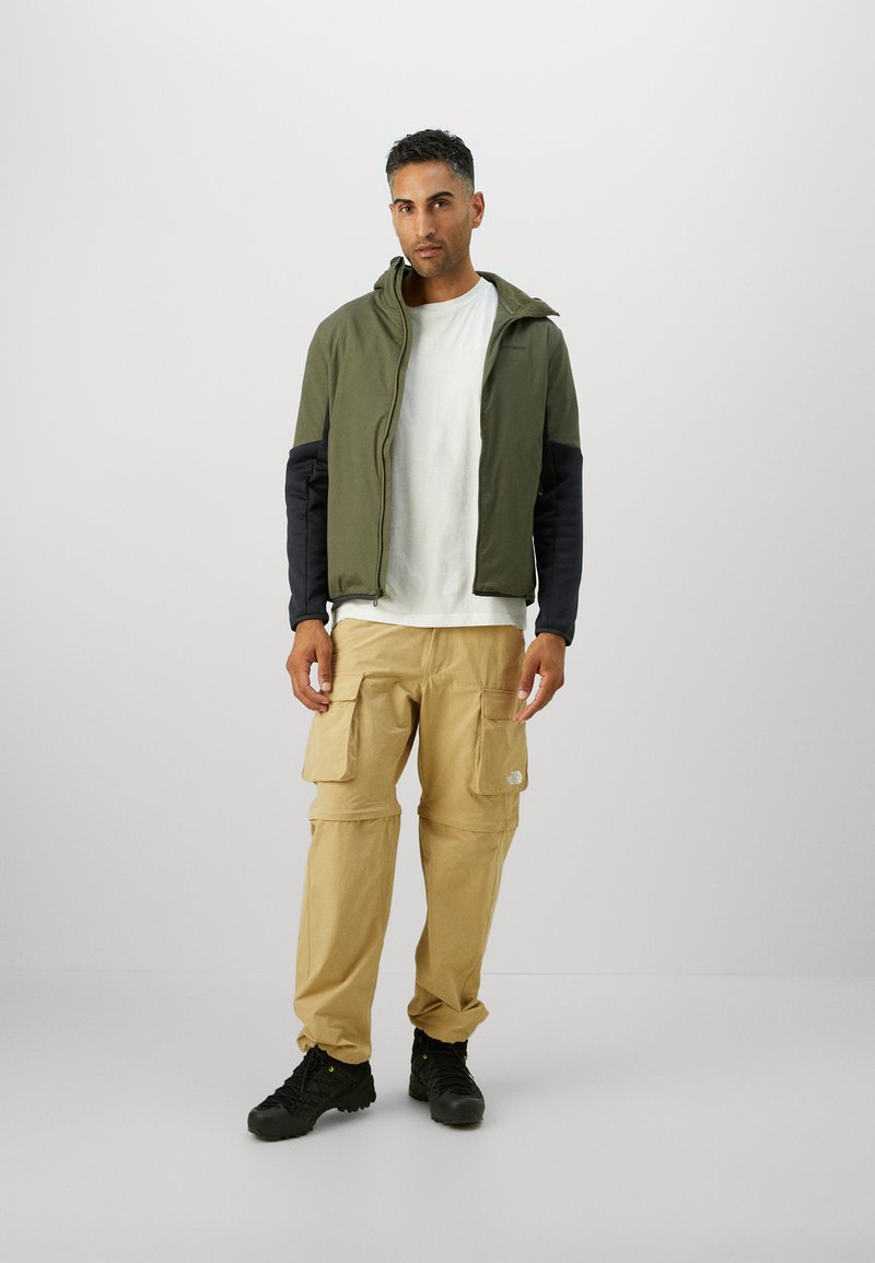 Oliven grøn jakke med marineblå ærmer, hvid t-shirt, beige cargo-bukser med lommer og sorte fodtøj, sat mod en ensfarvet baggrund.