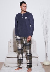 Granatowy, długi rękaw, pulower z dekoltem henley i logo; w zestawie z beżowymi i ciemnozielonymi spodniami w kratę flanelową. Model stoi boso.