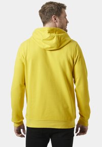 Sudadera con capucha amarilla hecha de tejido suave, con un bolsillo canguro, puños acanalados y un corte relajado. Se muestra desde la parte de atrás.