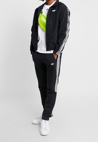 Svart lättviktsjacka med vita logodetaljer, parat med svarta sweatpants med vita sidostreck och en vit T-shirt. Ljusstark grön överkroppsväska.