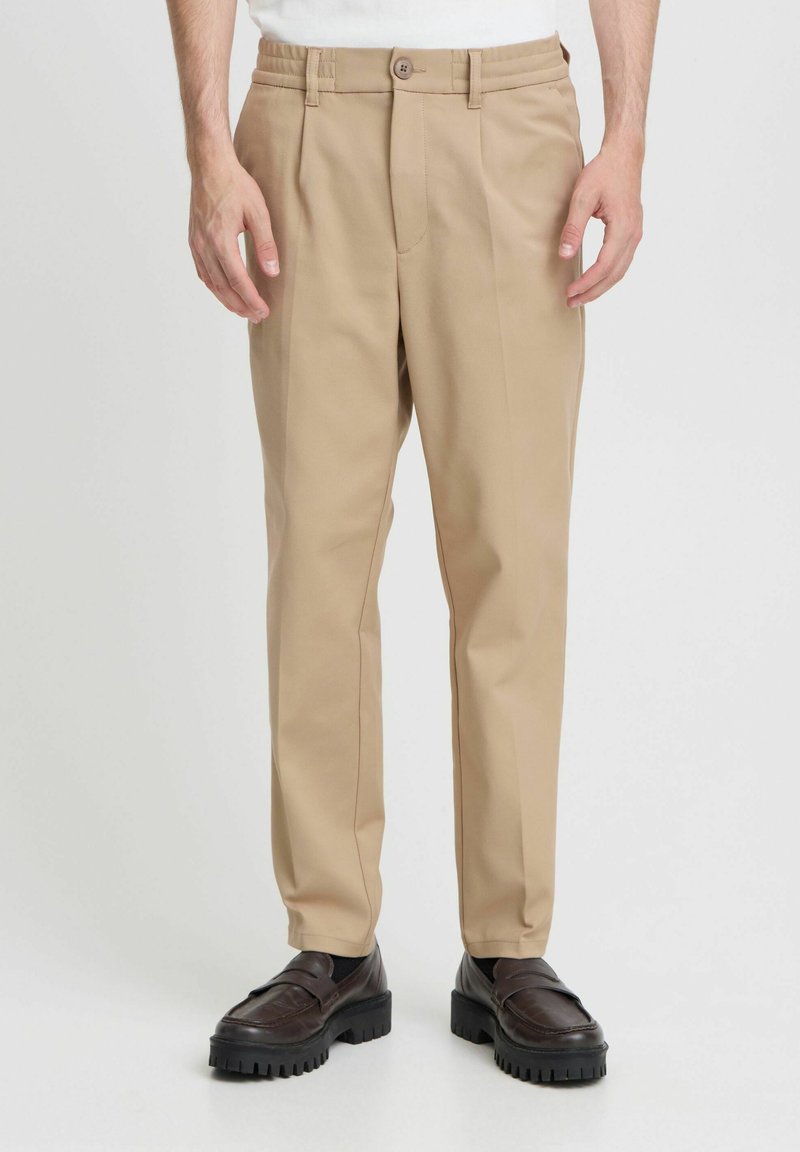 Beige tailored pants with a straight fit, belt loops, a button closure, and minimal creases. Worn with dark loafers. Smooth fabric texture.