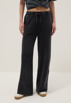 Person trägt gewaschene schwarze Sweatpants mit weitem Bein und Kordelzug an der Taille sowie weiße Sneakers mit Gummisohlen und steht vor einem schlichten Hintergrund.