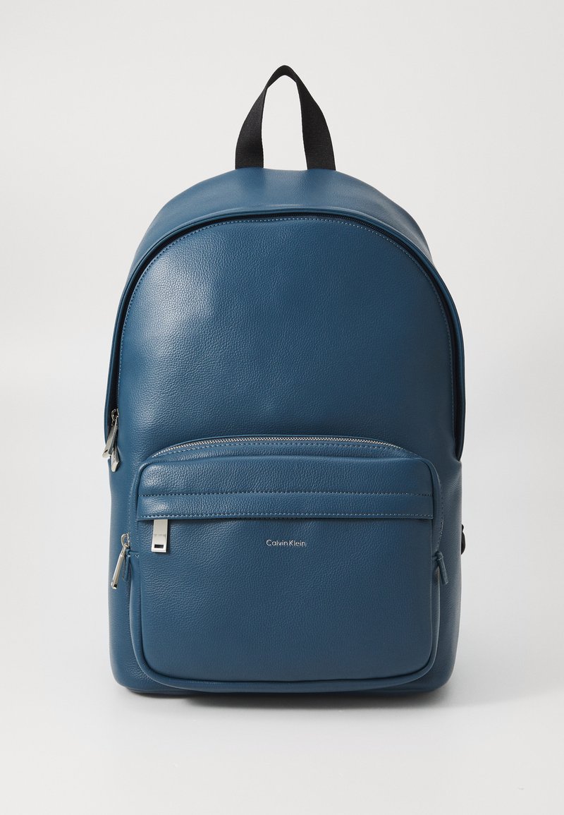 Mochila de cuero azul con forma redondeada, que cuenta con un bolsillo frontal con cremallera, herrajes plateados y un asa en la parte superior. Acentuada con un logotipo.