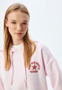 Sudadera rosa claro con cordón, que presenta un gráfico de una estrella roja y texto. Tejido suave, corte oversize, con una textura lisa y ribetes acanalados.