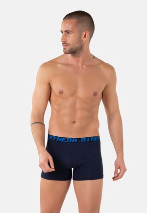 Homme torse nu aux cheveux courts regardant vers la gauche, portant un boxer bleu marine avec une ceinture bleue portant la mention "ATHENA".
