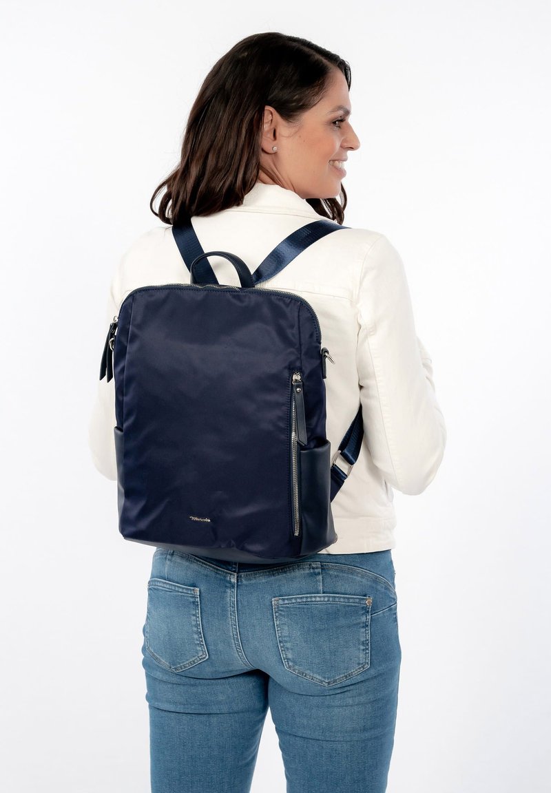 Mujer con una mochila azul oscuro con varios compartimentos con cremalleras, vestida con una chaqueta blanca y vaqueros azules, sonriendo y de perfil.