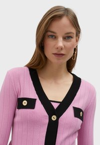 Rosa ribbad cardigan med svart detaljer, prydd med guldknappar och två bröstfickor. Designad med djup V-ringning och långa ärmar.