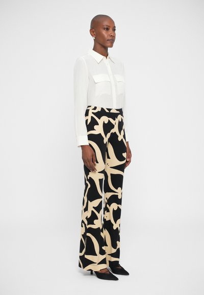 Camisa branca de botões com dois bolsos, combinada com calças de perna larga de cintura alta em um padrão abstrato em bege e preto. Sapatos pretos de bico fino.