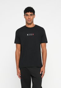 Μαύρο βαμβακερό T-shirt με στρογγυλή λαιμόκοψη, κοντά μανίκια και κεντρικό λογότυπο σε λευκό, κόκκινο και μπλε. Απλός, κλασικός σχεδιασμός.