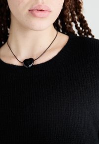 Suéter negro de punto con textura acanalada y un escote ceñido, combinado con un colgante negro en forma de corazón sobre un cordón delgado.