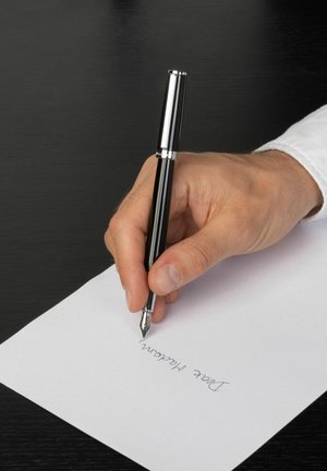 Penna stilografica nera con accenti argentati, tenuta in una mano mentre scrive su carta bianca. Sullo sfondo, una superficie di legno scuro.