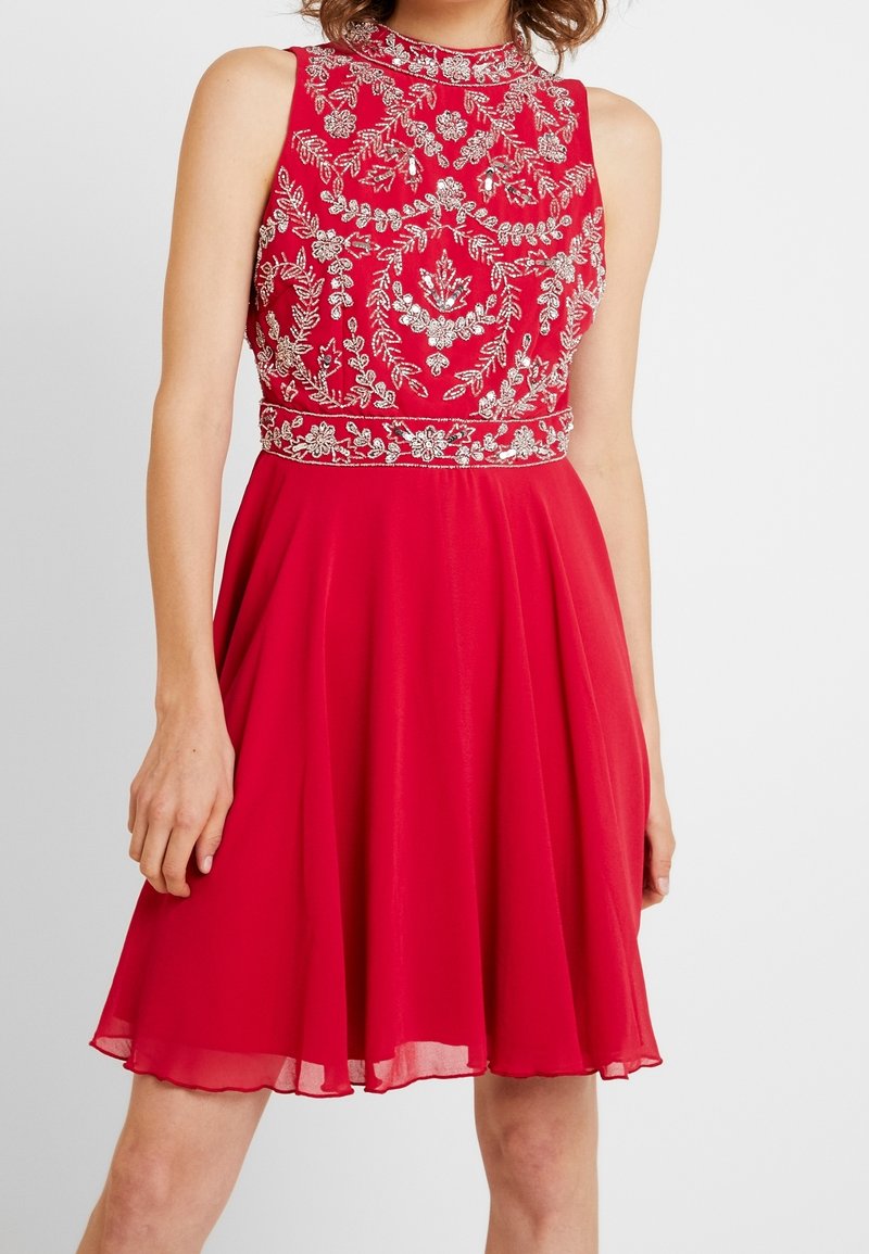 Robe rouge sans manches mi-longue avec un col montant et une broderie florale blanche sur le corsage et la ceinture, portée par une personne debout.