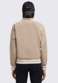 Beige bomberjack met witte strepen op de mouwen en onderrand, gemaakt van glad materiaal. Heeft een staande kraag en elastische manchetten, wat mooi past bij een zwarte pet.