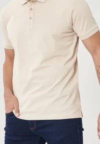 Beige polotröja med klassisk krage, tre knappar och ribbade ärmslut; tillverkad av ett slätt tyg, i kombination med mörkblå jeans.