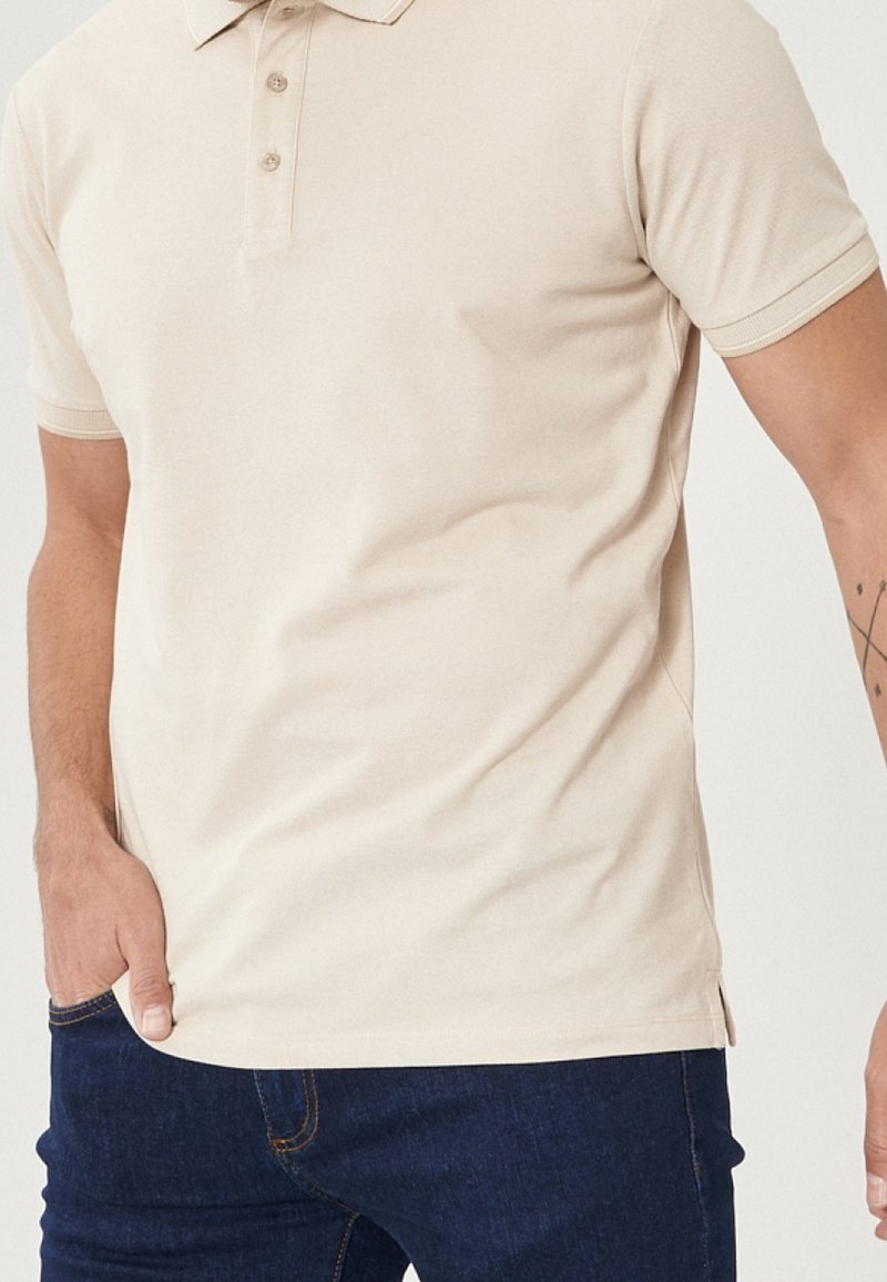 Beige polotröja med klassisk krage, tre knappar och ribbade ärmslut; tillverkad av ett slätt tyg, i kombination med mörkblå jeans.
