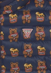 Tessuto di cotone blu navy trapuntato con orsetti di peluche ripetuti che stringono il logo GUESS e barattoli di miele etichettati "Miele", con una toppa centrale a forma di triangolo GUESS.
