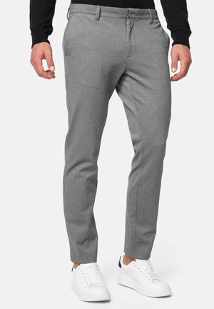Mann in schlanken grauen Hosen, schwarzem Langarmoberteil und weißen Sneakers, der vor einem einfarbigen hellen Hintergrund steht.