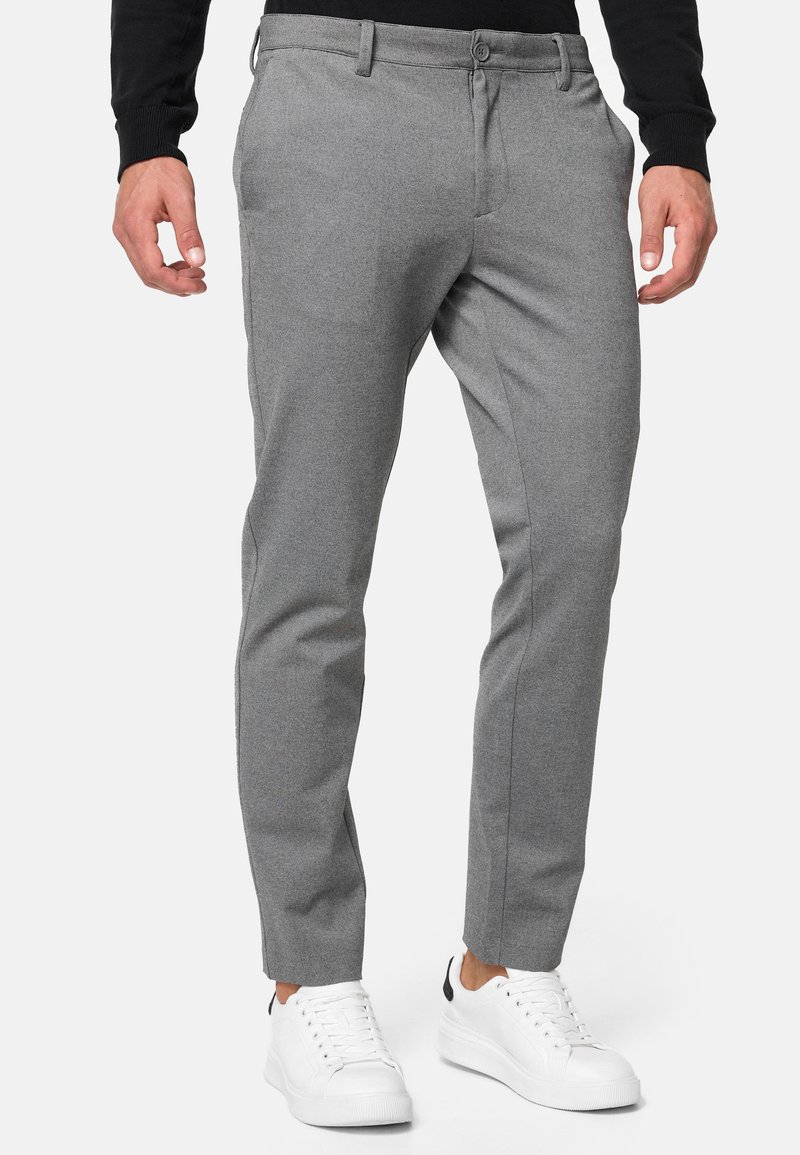 Mann iført slanke grå bukser, sort langermet topp og hvite sneakers, som står mot en ensfarget lys bakgrunn.