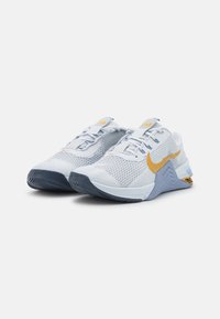 Bílé a šedé sportovní boty s textilním svrškem, s kultovním zlatým logem Nike a modrými detaily na podrážce a jazyku. Texturovaný design.