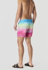Homme debout pieds nus portant un short de bain dégradé coloré et des sandales noires, vu de dos sur fond uni.