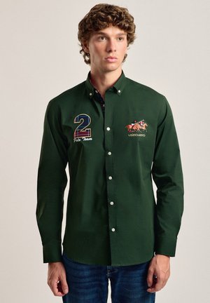 Camisa verde de manga larga, hecha de algodón, que presenta un número "2" en azul y un diseño ecuestre bordado en el pecho, con cuello abotonado.