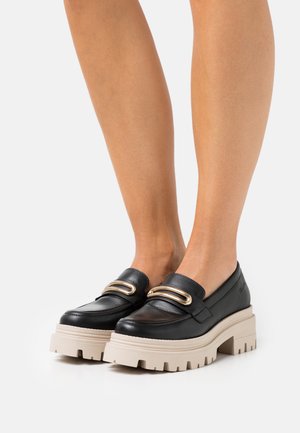 Mocassins en cuir noir avec une semelle plateforme beige épaisse, présentant des accents en métal doré et un design supérieur lisse. Texture douce.