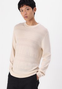 Bež pulover z dolgimi rokavi iz pletenine z strukturiranimi vodoravnimi vzorci in okroglim izrezom. Material izgleda mehak in lahek.