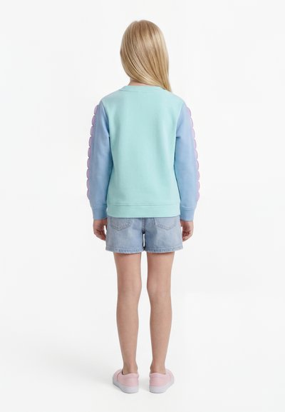 Enfant aux longs cheveux blonds portant un sweat-shirt vert menthe et bleu avec des accents de bulles roses sur les manches, un short en jean et des chaussures roses sans lacets.