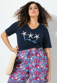 T-shirt di cotone blu scuro con scollatura a V e dettaglio annodato, con tre stelle marine bianche e la frase "Ho bisogno di vitamina mare". Pantaloni con motivo floreale.