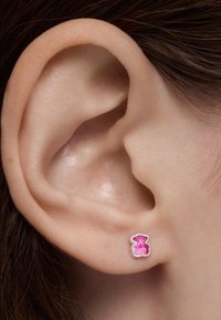 Primer plano de una oreja con un pequeño pendiente en forma de oso rosa en el lóbulo, cabello oscuro visible en el fondo.