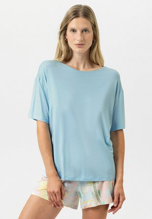 Donna con capelli biondi lunghi che indossa una maglietta azzurra a maniche corte e pantaloncini con motivo pastello, in piedi davanti a uno sfondo neutro.