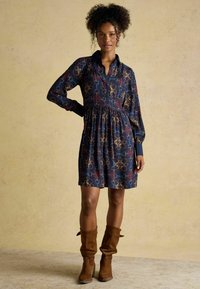 Merev navüüriga kleit pikkade varrukatega, volditud vöökoht ja mitmevärvilised geomeetrilised mustrid. Stylditud pruunide seemisnahast põlvikute kõrval.