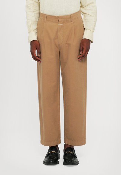 Beige Baumwollhosen mit lässiger Passform, die ein gerades Bein und einen dezenten Knick aufweisen. Getragen mit schwarzen Mokkasins, die eine glatte Textur zeigen.