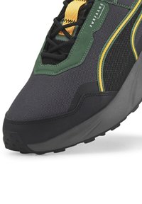 Nahaufnahme eines schwarzen und dunkelgrünen Sneakers mit gelben Akzenten, schwarzen Schnürsenkeln, strukturiertem Stoff und einer grauen, dicken Sohle.