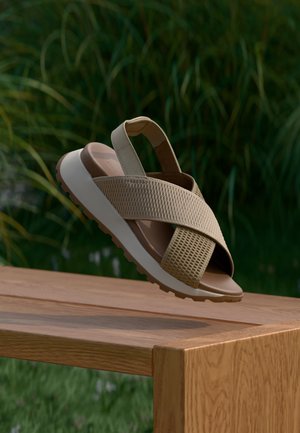 Sandalo beige con cinturini incrociati in mesh e suola bianca spessa, sospeso sopra una panchina di legno con sfondo sfocato di fogliame verde.