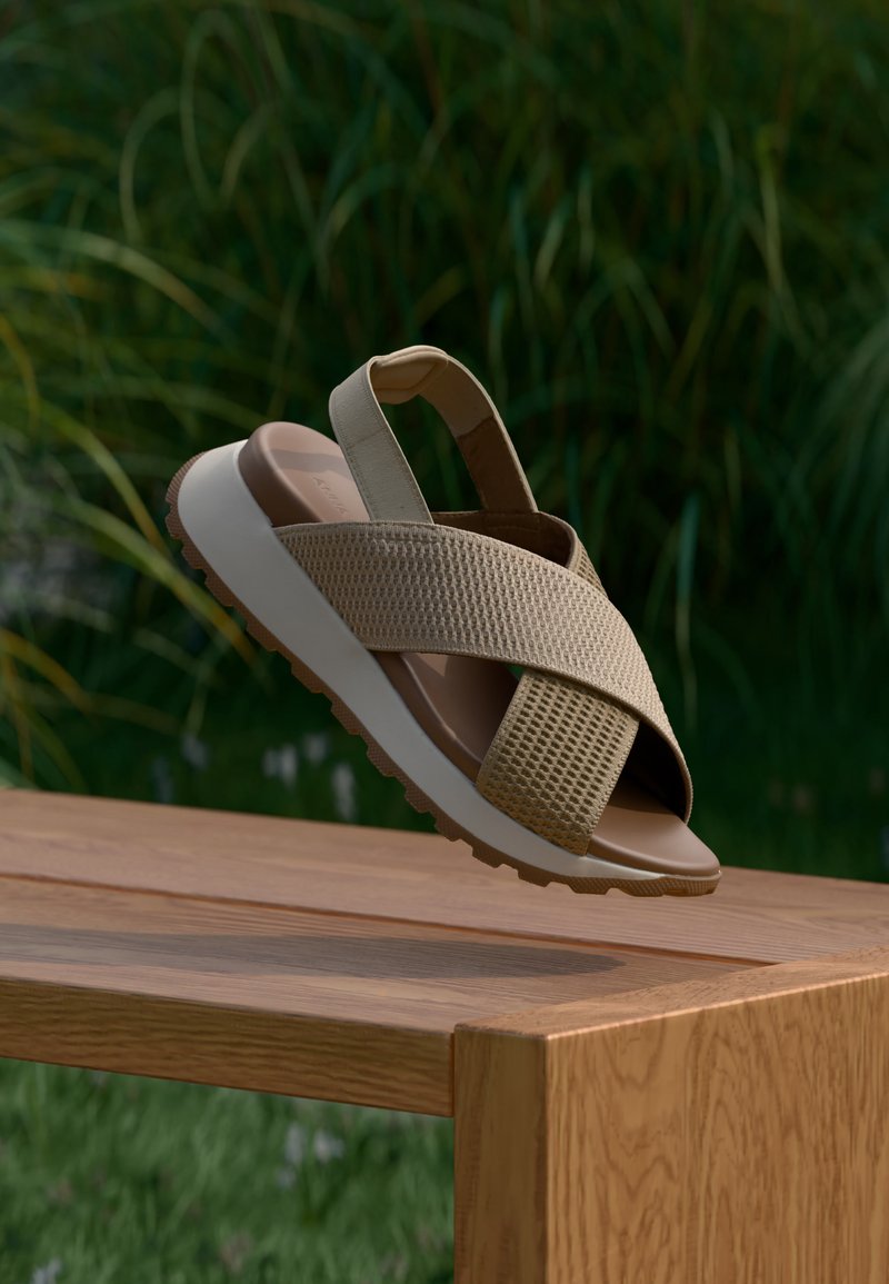 Sandale beige avec des brides en mesh croisées et une semelle épaisse blanche, flottant au-dessus d'un banc en bois avec un arrière-plan de feuillage vert flou.